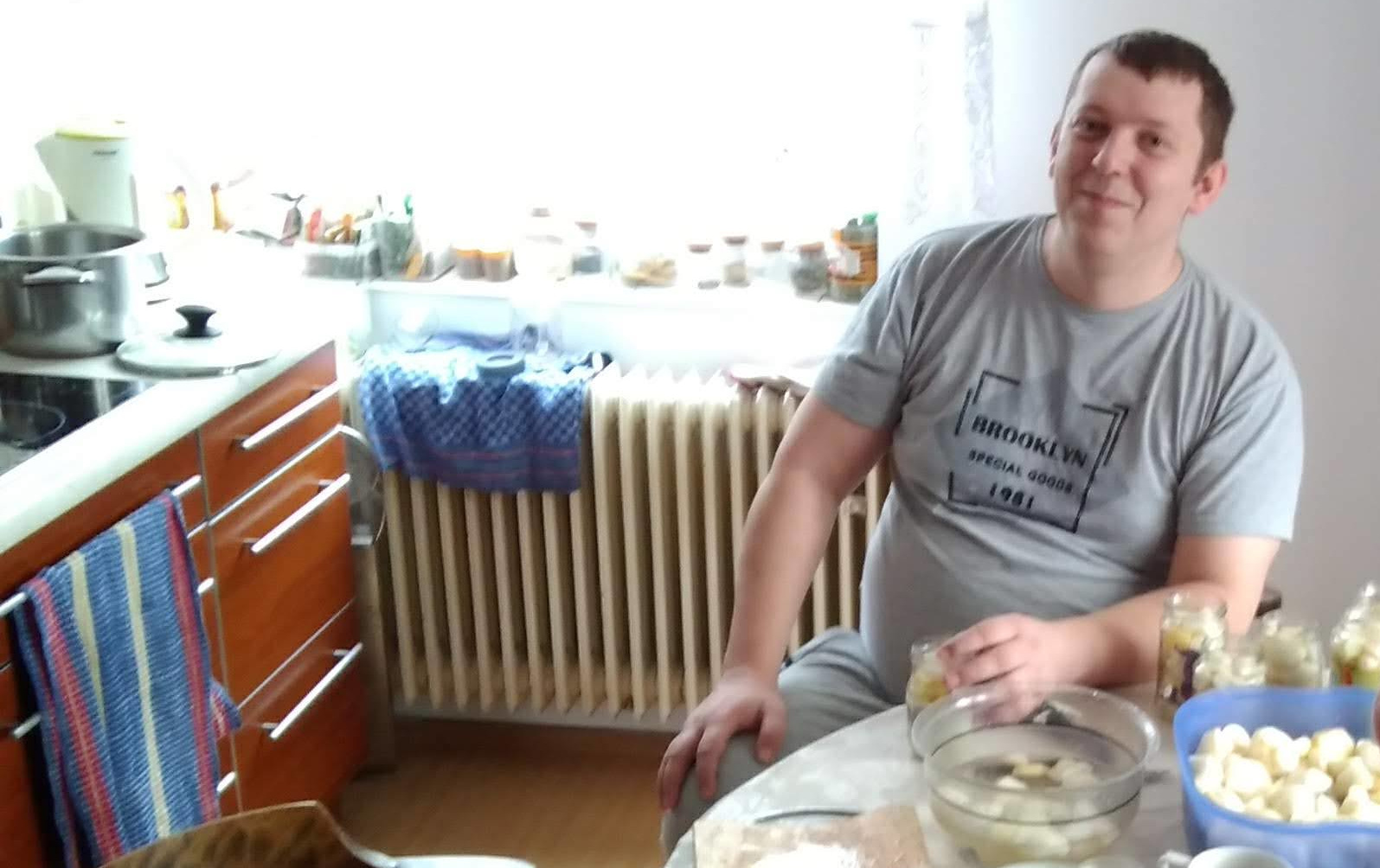 Jozef Môstka pri kuchynskom stole s misami so zeleninou pri zavaraní — fotografovaný bez varovania, usmieva sa.