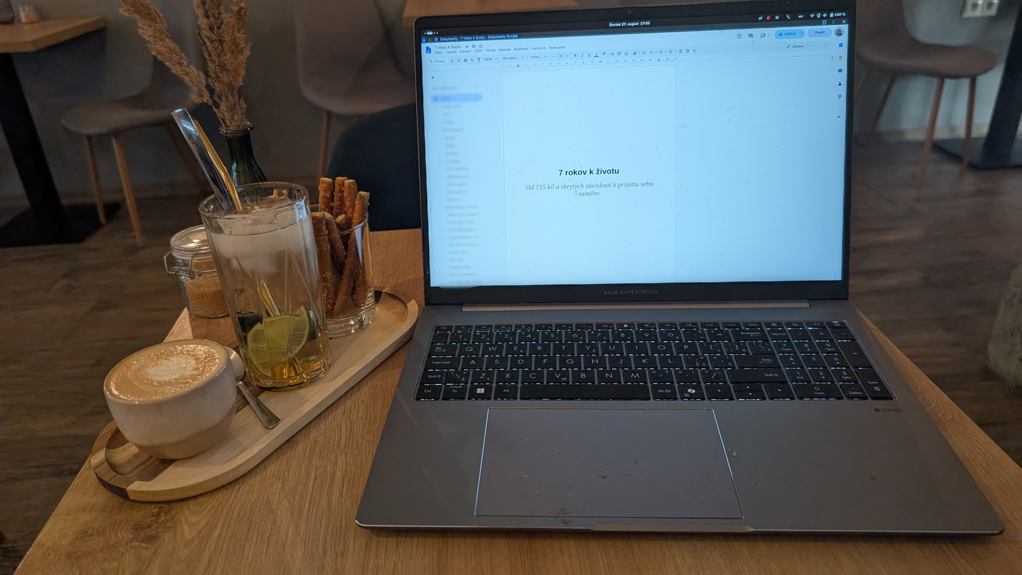 Otvorený notebook na drevenom stole v kaviarni, na obrazovke rukopis knihy 7 rokov k životu, vedľa káva a limonáda na podnose.