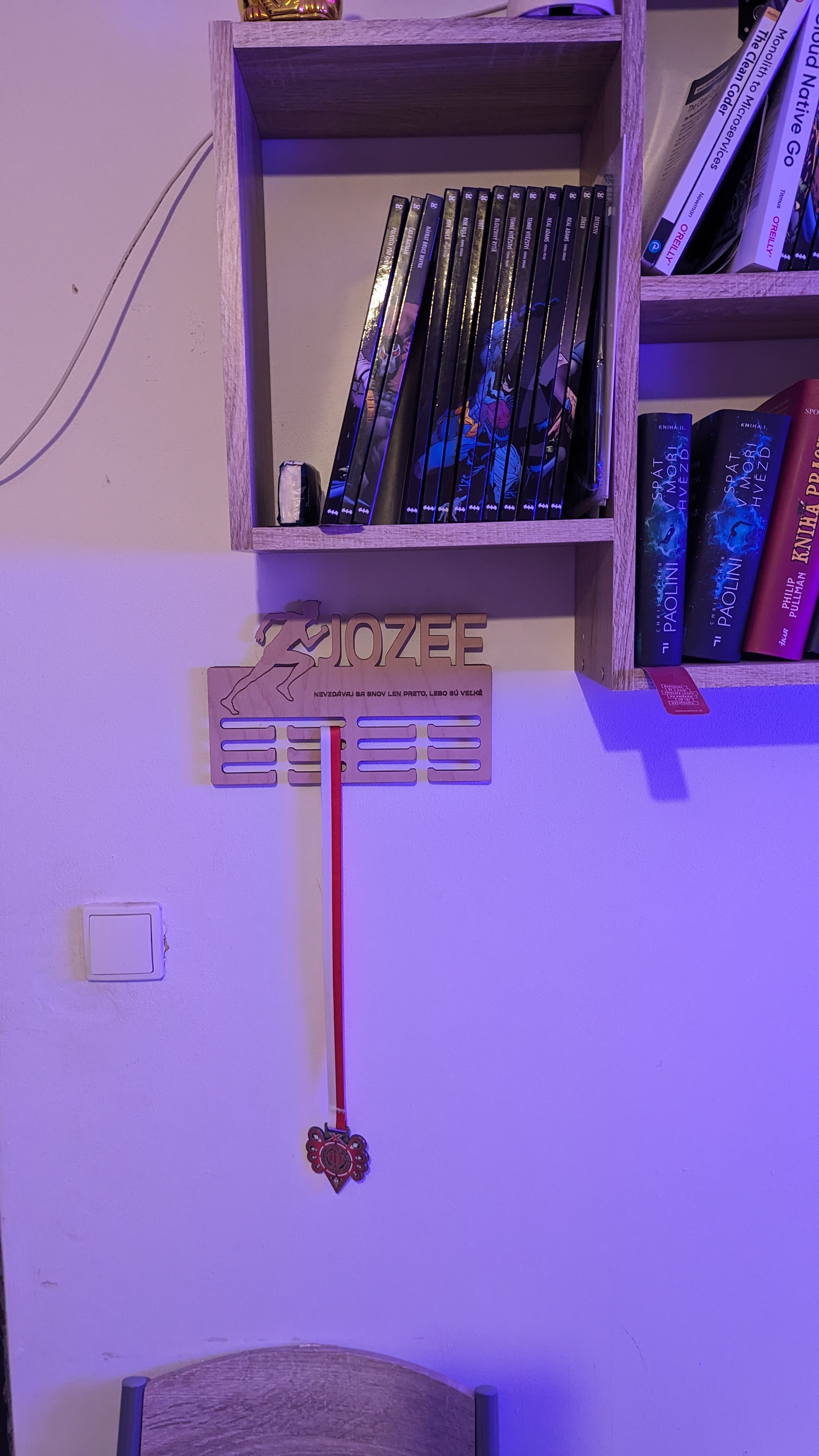 Drevený vešiak na medaily s nápisom JOZEE a vyrytým mottom, na ktorom visí jediná medaila na červenej stuhe.